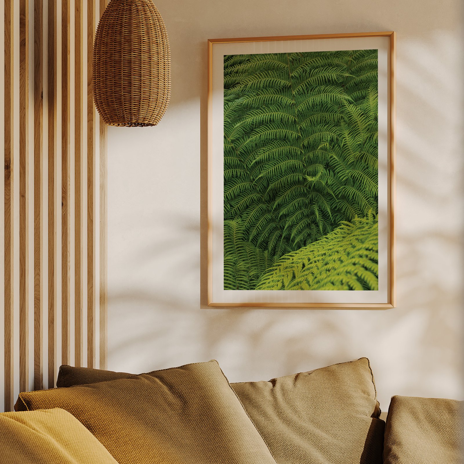 Vertical botanical close-up photograph of lush green fern fronds with intricate layering and texture in a natural wood frame, hung on a cream wall above a mustard-gold upholstered sofa, styled with wooden slat wall paneling and a woven rattan pendant light visible above.