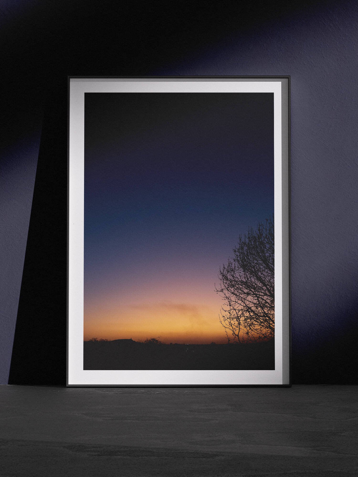 Vertical twilight landscape photograph showing a gradient sky transitioning from deep blue through purple tones to golden orange at the horizon, with a silhouetted bare tree branch on the right side, displayed in a white frame with white mat, leaning against a dark navy wall in a gallery-style floor presentation with dramatic side lighting.