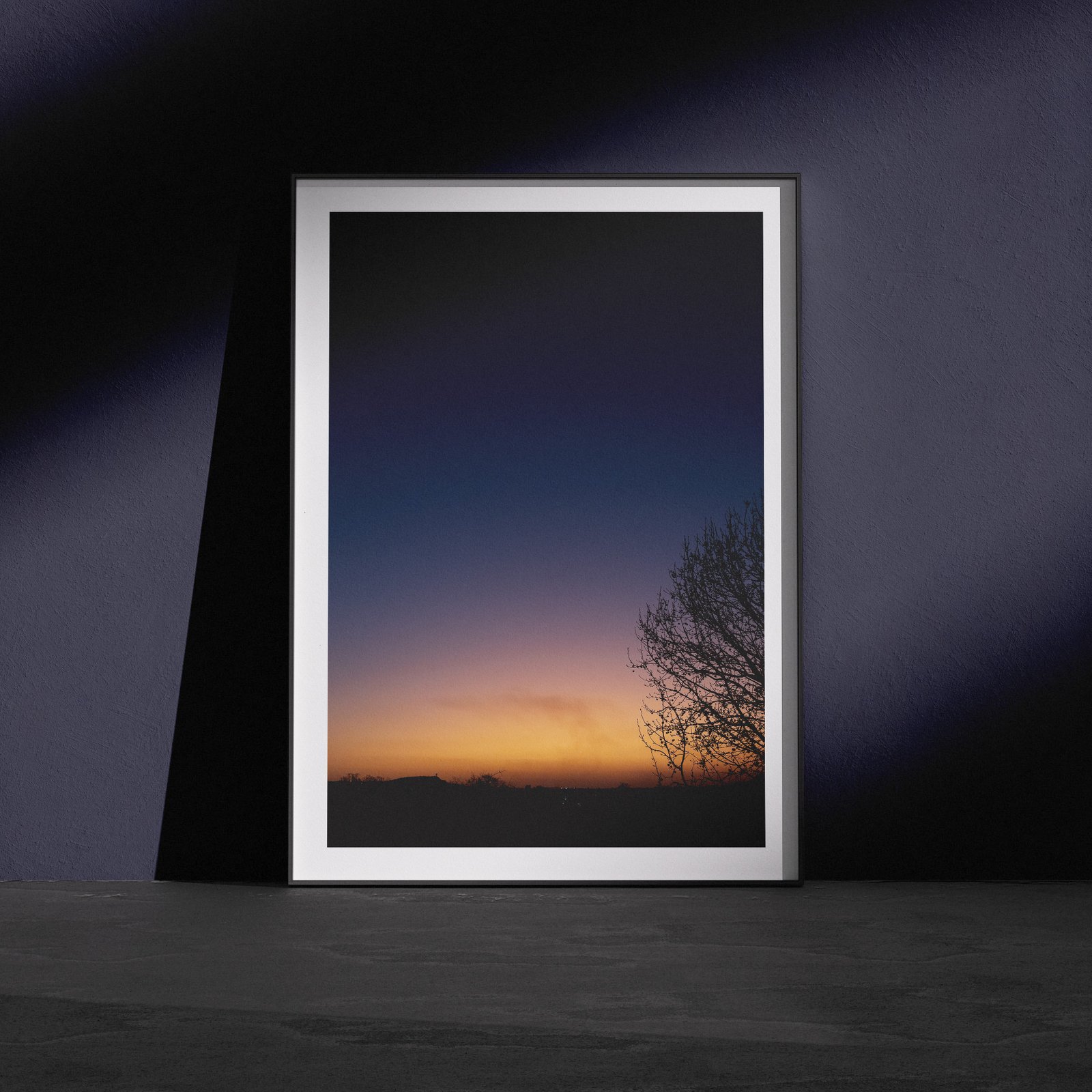 Vertical twilight landscape photograph showing a gradient sky transitioning from deep blue through purple tones to golden orange at the horizon, with a silhouetted bare tree branch on the right side, displayed in a white frame with white mat, leaning against a dark navy wall in a gallery-style floor presentation with dramatic side lighting.