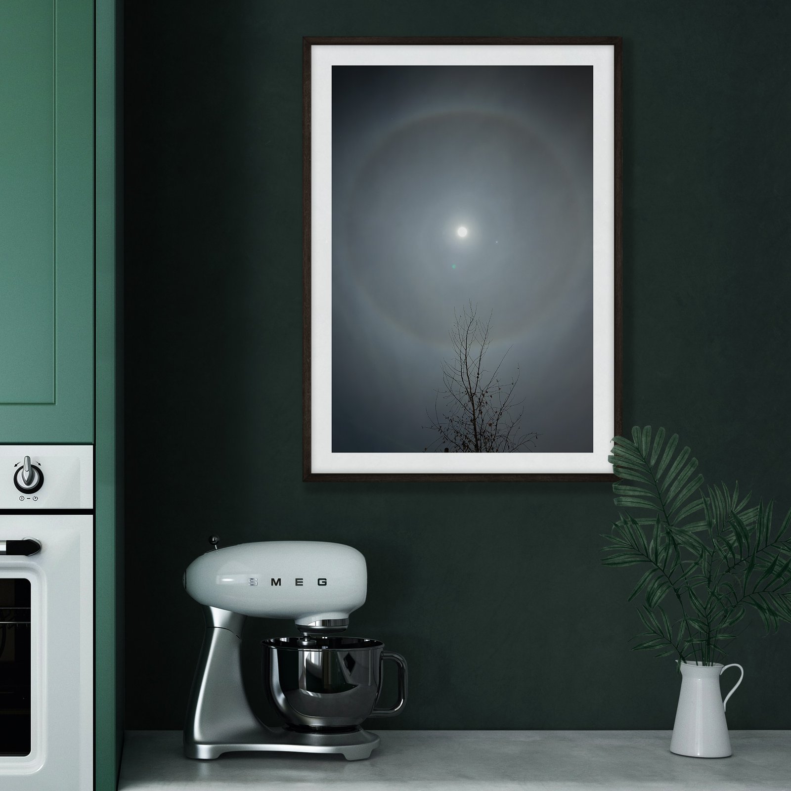 Vertical nocturnal sky photograph showing a glowing full moon and silhouetted bare branch in a white frame, hung on a rich dark green kitchen wall above a vintage cream and black retro coffee maker (SMEG style), styled with a white ceramic pitcher.