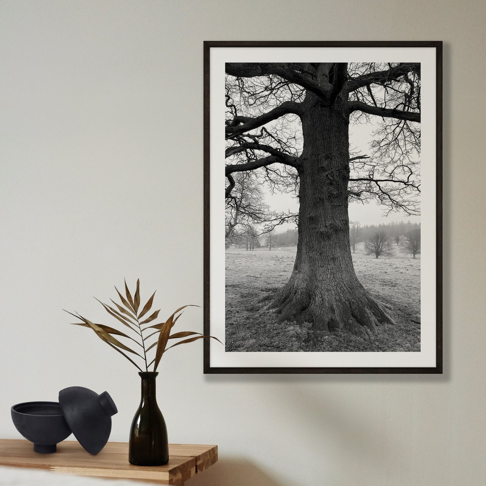 Vertical black and white landscape photograph of a majestic solitary oak tree with bare winter branches standing in a misty field with distant landscape, displayed in a black frame with white mat on a pale beige wall, styled with a minimalist shelf arrangement below featuring a black vase with dried grasses and black ceramic bowl.